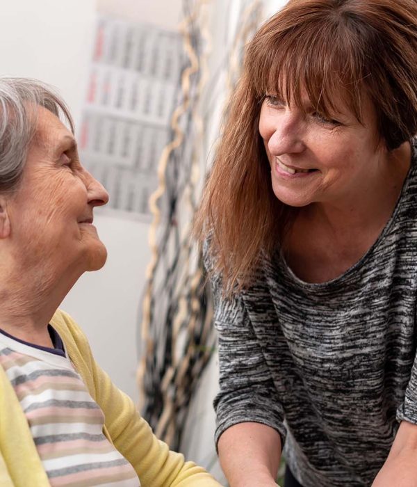 Aidante à domicile souriante échangeant avec une personne âgée assise, moment chaleureux d’accompagnement et de soutien.