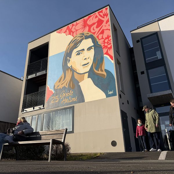 Fresque murale représentant une figure féminine engagée sur la façade d’un immeuble urbain avec des passants au premier plan