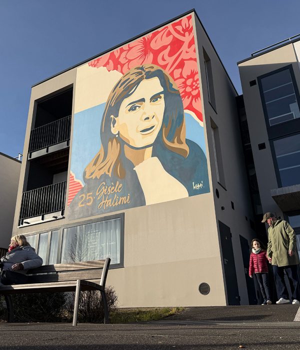 Fresque murale représentant une figure féminine engagée sur la façade d’un immeuble urbain avec des passants au premier plan