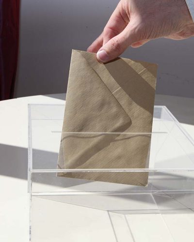 Main déposant un bulletin de vote dans une urne transparente, symbolisant participation citoyenne et processus électoral.