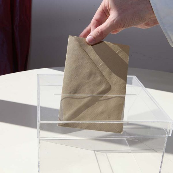 Main déposant un bulletin de vote dans une urne transparente, symbolisant participation citoyenne et processus électoral.